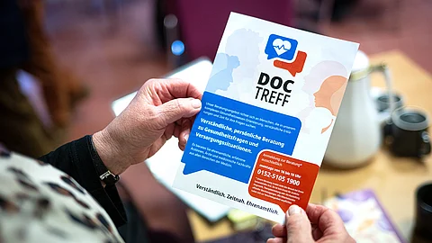 Eine Person hält einen Flyer mit der Aufschrift „Doc Treff“ in den Händen; darauf sind stilisierte Gesichter und Informationen zu einem medizinischen Beratungsangebot zu sehen.