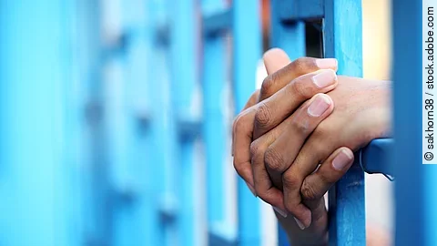 Hände eines Gefangenen, die sich zwischen blauen Gitterstäben verschränken