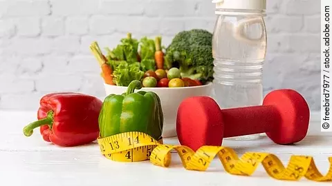 Frisches Gemüse wie Paprika und Zucchini liegt neben einer roten Hantel auf einem hellen Untergrund. 