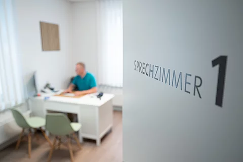 Ein Hausarzt sitzt in einem Sprechzimmer seiner Hausarztpraxis an einem Schreibtisch und arbeitet am Computer.