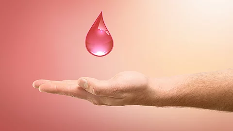 Eine ausgestreckte Hand hält einen roten Blutstropfen vor einem sanften rosa Hintergrund