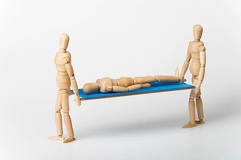 Zwei Holzfiguren tragen eine dritte liegende Figur auf einer Trage mit blauer Fläche vor neutralem Hintergrund. Zwei Holzfiguren tragen eine dritte liegende Figur auf einer Trage mit blauer Fläche vor neutralem Hintergrund.