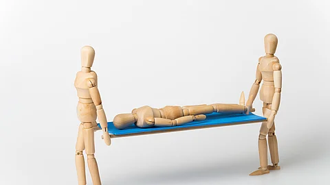 Zwei Holzfiguren tragen eine dritte liegende Figur auf einer Trage mit blauer Fläche vor neutralem Hintergrund.
