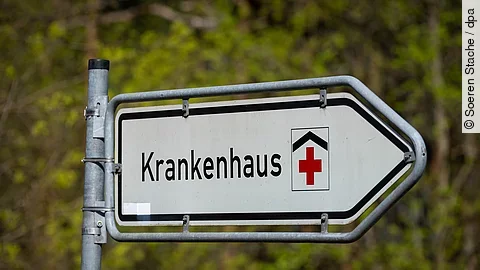 Straßenschild mit der Aufschrift ‚Krankenhaus‘ und rotem Kreuz, das auf eine Klinik hinweist. 