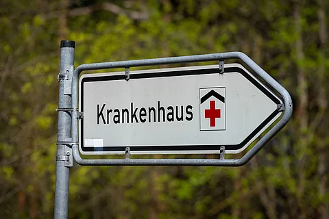 Straßenschild mit der Aufschrift ‚Krankenhaus‘ und rotem Kreuz, das auf eine Klinik hinweist. 