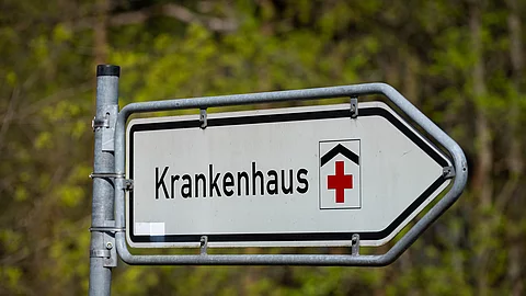 Straßenschild mit der Aufschrift ‚Krankenhaus‘ und rotem Kreuz, das auf eine Klinik hinweist. 