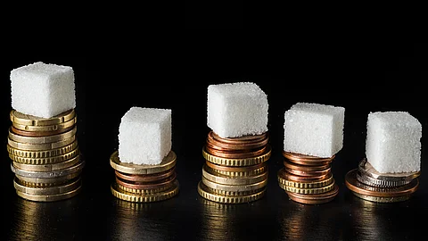 Mehrere Würfelzucker stehen jeweils auf unterschiedlich hohen Stapeln aus Euro‑Münzen vor schwarzem Hintergrund