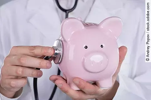 Ein Arzt hält ein rosafarbenes Sparschwein in den Händen und hört es mit einem Stethoskop ab.