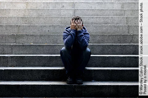 ein junger Mann sitzt auf einer Betontreppe und hält die Hände vor das Gesicht