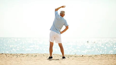 Ein älterer Herr in Sportkleidung steht am Strand und führt eine seitliche Dehnübung aus, mit Meer und hellem Himmel im Hintergrund.