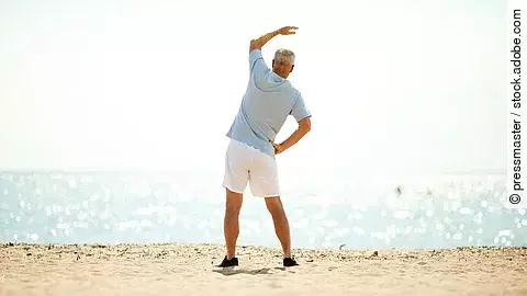 Ein älterer Herr in Sportkleidung steht am Strand und führt eine seitliche Dehnübung aus, mit Meer und hellem Himmel im Hintergrund.