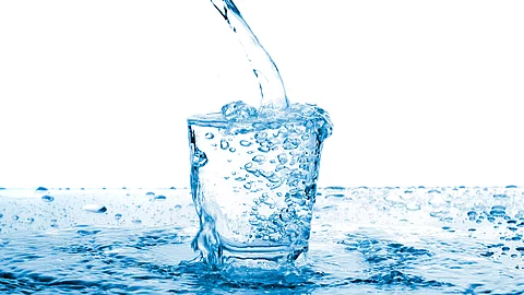 Ein Glas klares Wasser, in das von oben Wasser eingegossen wird; umgeben von spritzenden Tropfen 