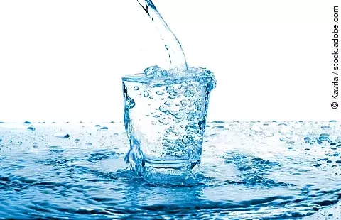 Ein Glas klares Wasser, in das von oben Wasser eingegossen wird; umgeben von spritzenden Tropfen 