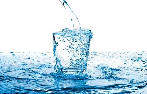 Ein Glas klares Wasser, in das von oben Wasser eingegossen wird; umgeben von spritzenden Tropfen 