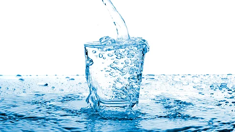 Ein klarer Wasserstrahl fließt in ein Glas und erzeugt sprudelnde Blasen; das Glas steht in einer Wasserfläche