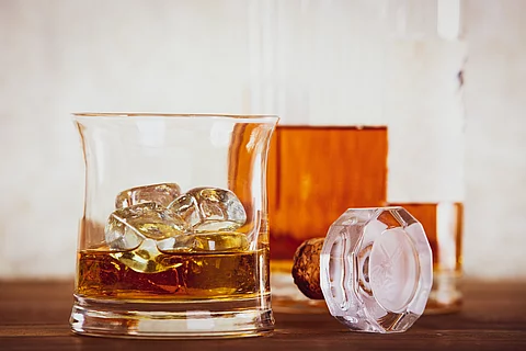 Nahaufnahme eines Whiskyglases mit Eiswürfeln, daneben eine geöffnete Flasche Whisky und ein Glasverschluss auf einem Holztisch.