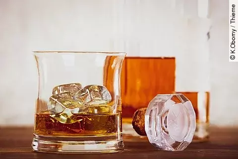 Nahaufnahme eines Whiskyglases mit Eiswürfeln, daneben eine geöffnete Flasche Whisky und ein Glasverschluss auf einem Holztisch.
