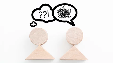 Sprachbarrieren: Zwei symbolische Holzfiguren stehen sich gegenüber, über ihren Köpfen Sprechblasen mit Fragezeichen und unverständlichen Zeichen
