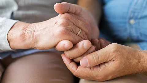 Nahaufnahme zweier Erwachsener, die nebeneinandersitzen und ihre Hände auf einem Knie ineinandergelegt halten Nahaufnahme zweier Erwachsener, die nebeneinandersitzen und ihre Hände auf einem Knie ineinandergelegt halten