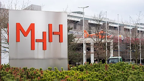 Das große Eingangsschild der Medizinischen Hochschule Hannover mit roten Buchstaben „MHH“, davor Sträucher, dahinter ein Klinikgebäude und ein Rettungshubschrauber auf einer Plattform.