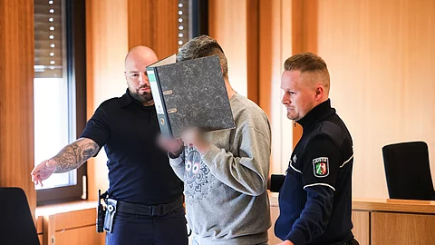 Ein Angeklagter hält einen Aktenordner vor sein Gesicht, während er von zwei Justizbeamten in einen Gerichtssaal geführt wird.
