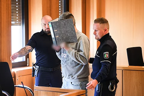 Ein Angeklagter hält einen Aktenordner vor sein Gesicht, während er von zwei Justizbeamten in einen Gerichtssaal geführt wird.
