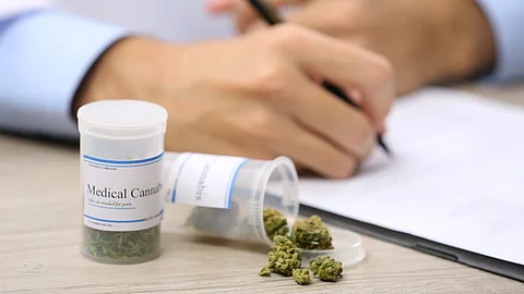 Eine ärztliche Hand schreibt ein Rezept auf einem Klemmbrett; daneben liegen medizinische Cannabisblüten, eine Pipette und mehrere Medikamentenfläschchen auf einem Tisch.