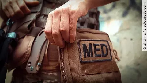 Nahaufnahme einer Hand, die eine braune taktische Sanitätstasche mit der Aufschrift „MED“ öffnet; daneben Ausrüstung wie ein Klappmesser.