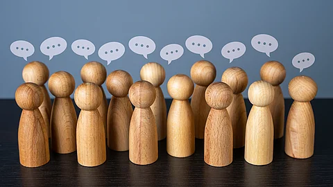 Mehrere stilisierte Holzfiguren stehen dicht beieinander, über ihren Köpfen befinden sich Sprechblasen
