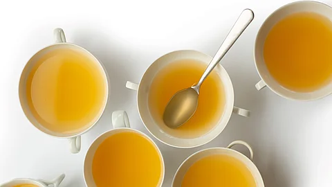 Mehrere weiße Tassen mit klarer, gelblicher Gemüsebrühe stehen von oben fotografiert auf einem hellen Hintergrund, in einer Tasse liegt ein Löffel.