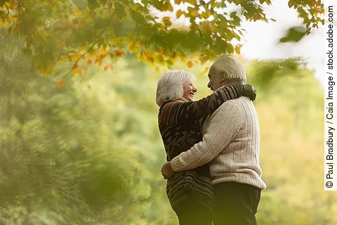 Zwei ältere Personen umarmen sich in einer herbstlichen Parklandschaft, umgeben von grünem und gelbem Laub. Zwei ältere Personen umarmen sich in einer herbstlichen Parklandschaft, umgeben von grünem und gelbem Laub.