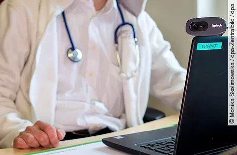 Ein Arzt in weißem Kittel mit Stethoskop sitzt vor einem Laptop mit Webcam.