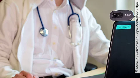 Ein Arzt in weißem Kittel mit Stethoskop sitzt vor einem Laptop mit Webcam.