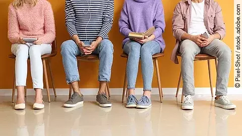 Vier Personen sitzen nebeneinander auf Holzstühlen im Wartebereich einer Arztpraxis.