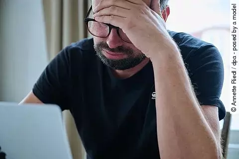 Ein Mann sitzt an einem Tisch vor einem Laptop, hält sich den Kopf und seufzt.