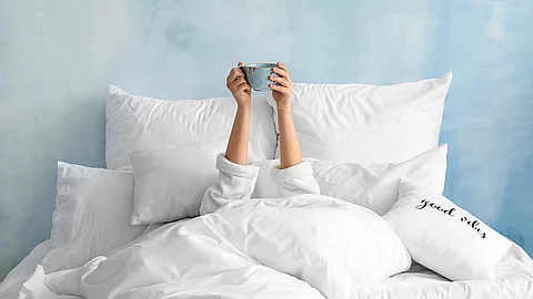 Eine Person liegt vollständig in weißer Bettwäsche, ist bis auf die Arme unter Decken verborgen und hält eine Tasse über dem Kopf; im Hintergrund eine hellblaue Wand und ein Kissen mit der Aufschrift „good vibes“.