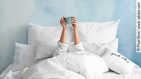 Eine Person liegt vollständig in weißer Bettwäsche, ist bis auf die Arme unter Decken verborgen und hält eine Tasse über dem Kopf; im Hintergrund eine hellblaue Wand und ein Kissen mit der Aufschrift „good vibes“.
