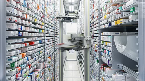 Ein automatisiertes Lagersystem mit hohen, dicht bestückten Regalen, in denen zahlreiche Medikamentenpackungen lagern; im Zentrum fährt ein Roboterarm auf einer vertikalen Schiene.