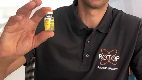 Hand hält ein kleines Glasfläschchen mit gelbem Warnetikett für radioaktives Material – darin wird ein Radiopharmakon für diagnostische oder therapeutische Zwecke abgefüllt.