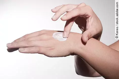 Nahaufnahme einer Hand, auf deren Handrücken eine weiße Creme aufgetragen wird. Eine zweite Hand verteilt die Creme sanft auf der Haut.