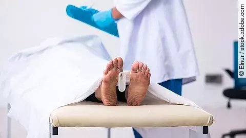 Ein medizinischer Fachmann steht neben einer Liege, auf der ein zugedeckter Körper mit sichtbaren Füßen liegt. An einem Fuß hängt ein Identifikationsanhänger.