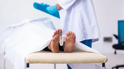 Ein medizinischer Fachmann steht neben einer Liege, auf der ein zugedeckter Körper mit sichtbaren Füßen liegt. An einem Fuß hängt ein Identifikationsanhänger.