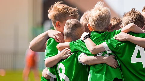 Kinder in grünen Sporttrikots stehen eng beisammen auf einem Sportplatz und legen im Team die Arme umeinander.
