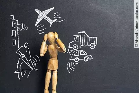 Eine Holz-Gliederpuppe vor einer dunklen Tafeloberfläche mit verschiedenen Symbolen: ein Flugzeug, ein Auto, ein Lastwagen und ein Presslufthammer.