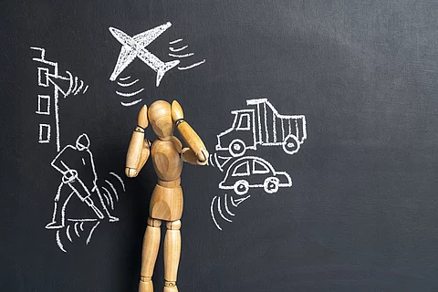 Eine Holz-Gliederpuppe vor einer dunklen Tafeloberfläche mit verschiedenen Symbolen: ein Flugzeug, ein Auto, ein Lastwagen und ein Presslufthammer.
