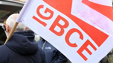 Eine Person hält eine weiße Fahne mit großen roten Buchstaben „IG BCE“; im Hintergrund sind weitere Personen und eine Außenszene erkennbar.