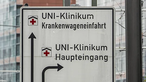 Ein schwarz-weißes Hinweisschild an einem Metallpfosten zeigt eine Richtungsangabe zum Haupteingang und zur Krankenwageneinfahrt des Uniklinikums Frankfurt