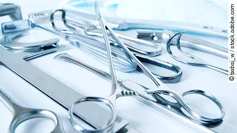 Auf einer hellen Unterlage liegen verschiedene chirurgische Instrumente wie Klemmen, Skalpelle, Scheren und metallische Halterungen, unsortiert und teilweise überlappend angeordnet