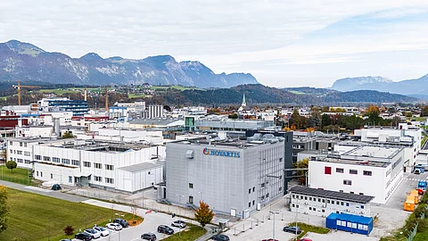 Luftaufnahme eines großen Pharmaproduktionsstandorts mit mehreren Industriegebäuden vor alpiner Landschaft – Standort des Konzerns Novartis in Kundl, Österreich.