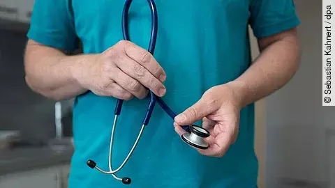 Ein Hausarzt steht in seiner Hausarztpraxis und hält ein Stethoskop. 